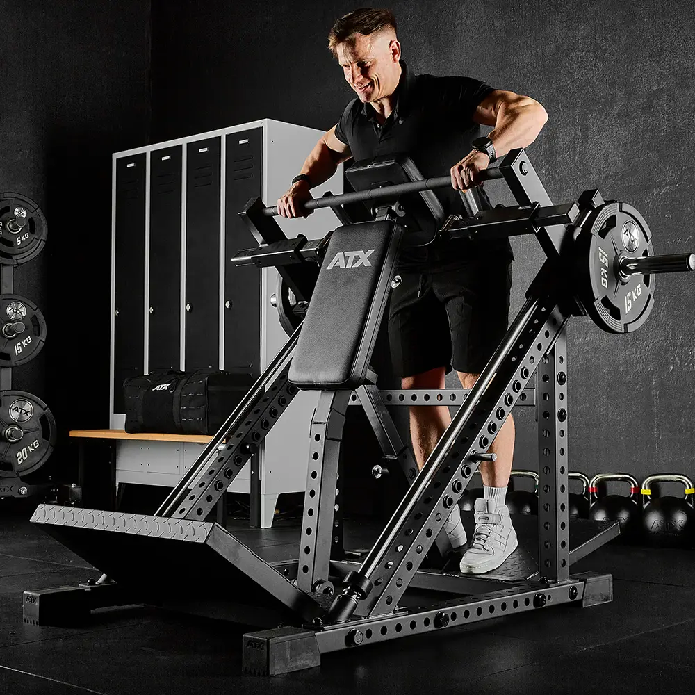 A person in gym attire uses a black ATX leverage squat machine in a gym with kettlebells, weight plates, an ATX® Smith Row Machine, and lockers visible in the background.
