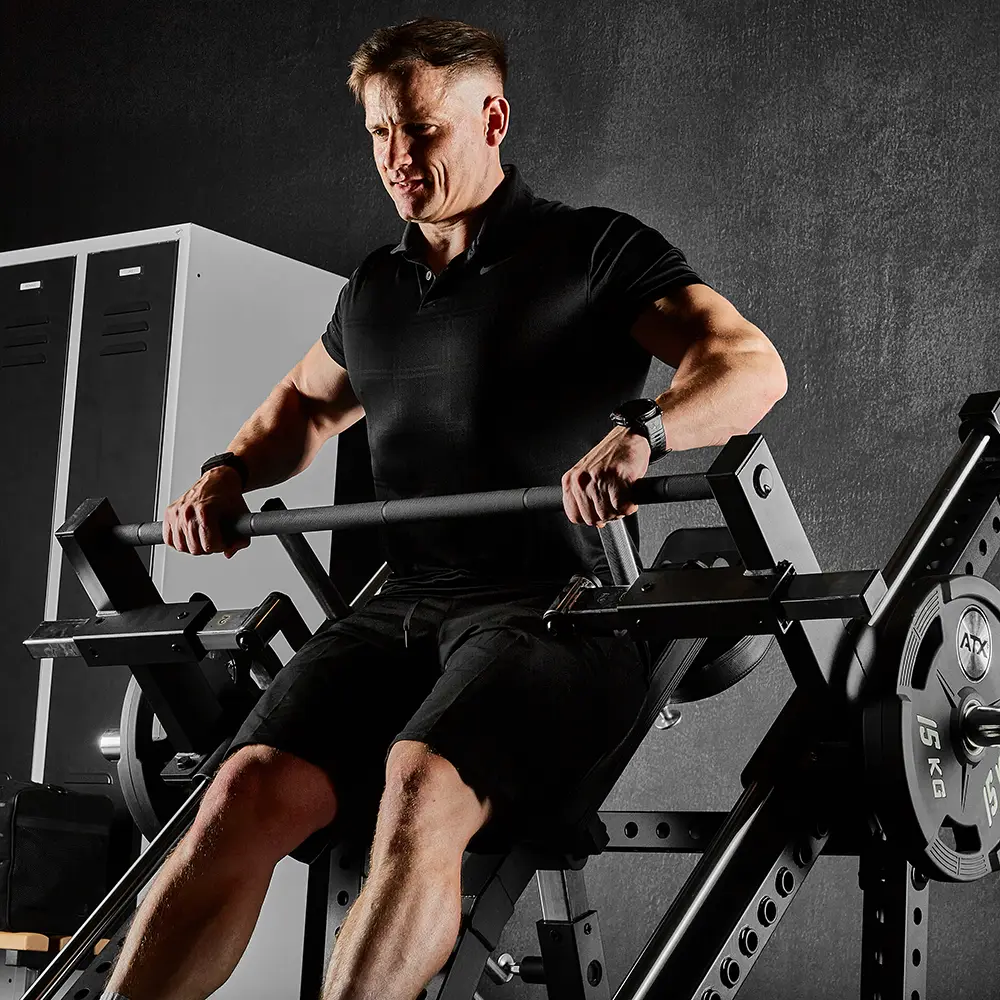 A man in black workout clothes uses a leg press machine and the ATX® Smith Row Machine in a gym, gripping the handles and pushing weights, with lockers visible in the background.