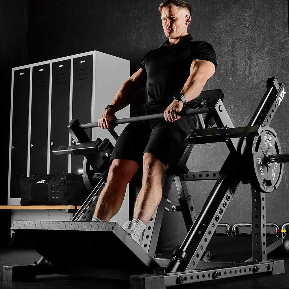 A man in black gym clothes uses a lever squat machine with weights in a gym, pushing up with his legs. In the background, among the black lockers and other equipment, an ATX® Smith Row Machine is visible.