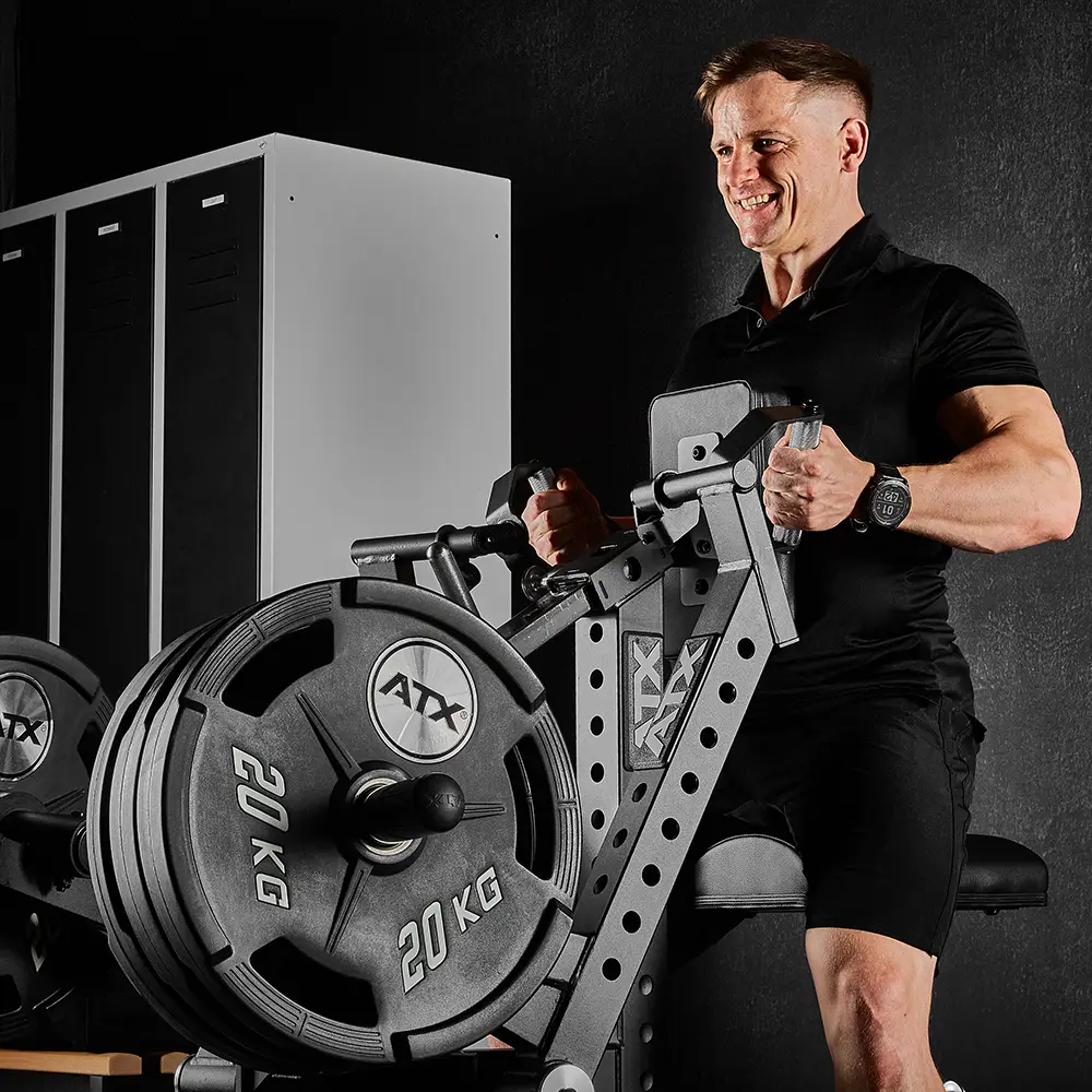 A man in black workout clothes uses an ATX® Chest Supported Row Machine loaded with multiple 20 kg weight plates in a gym, with lockers visible in the background.