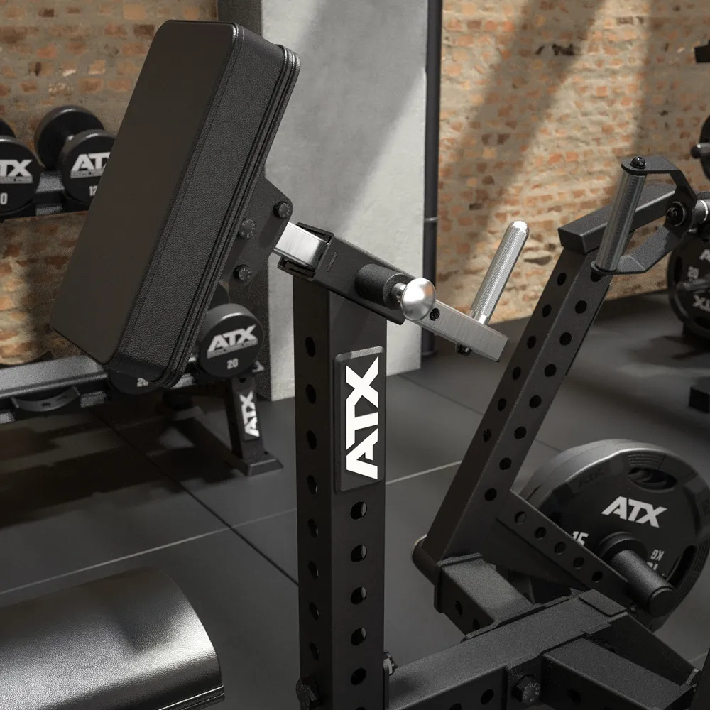 A close-up of a black ATX gym preacher curl bench in a weightlifting area, with dumbbells, weight plates, and a brick wall visible in the background.