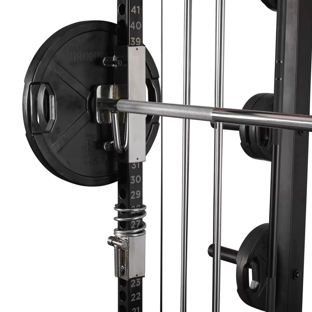 Close-up of a gym machine showing a barbell loaded with a 45 lb (20.4 kg) weight plate, attached to a rack with numbered height settings and an adjustable safety latch. Additional weight plates are stored in the background.