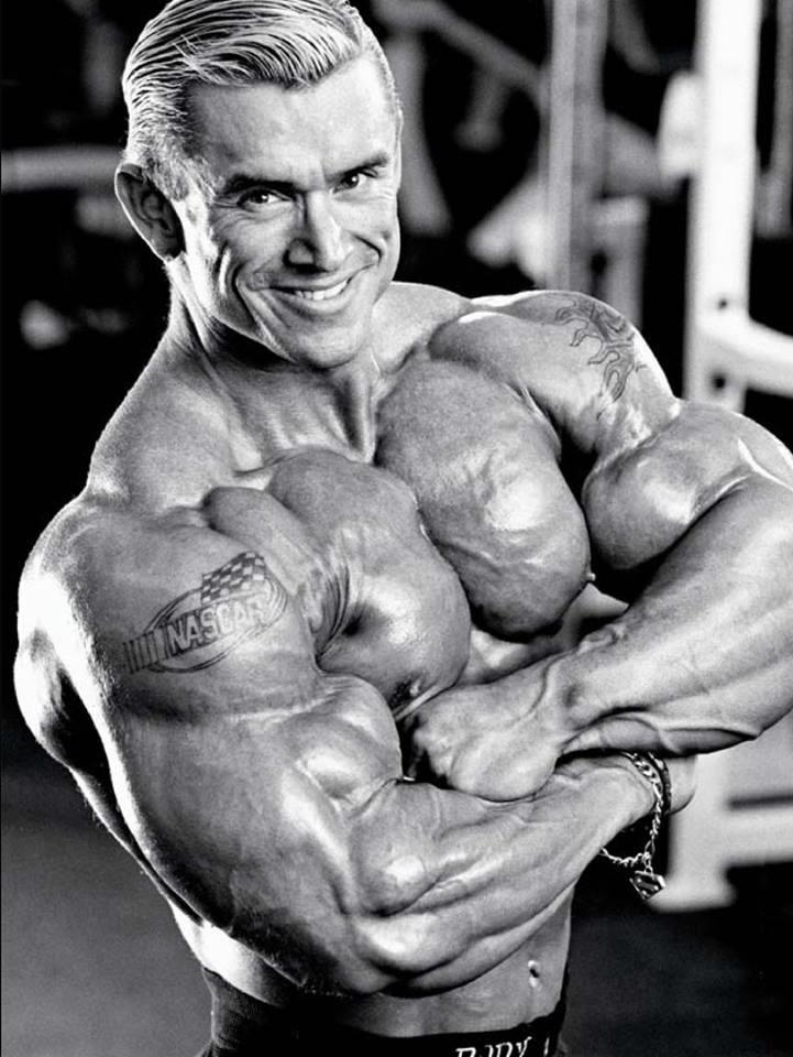 A muscular man with slicked-back hair poses in a gym, showing off his flexed arms and upper body. He is shirtless, smiling, and has visible tattoos, including a NASCAR logo on his upper arm. The image is in black and white.