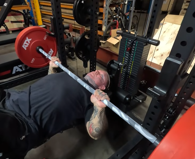 A man with tattoos lies on a bench press bench in a gym, gripping a barbell with red weight plates, preparing to lift. Gym equipment and weights are visible in the background.