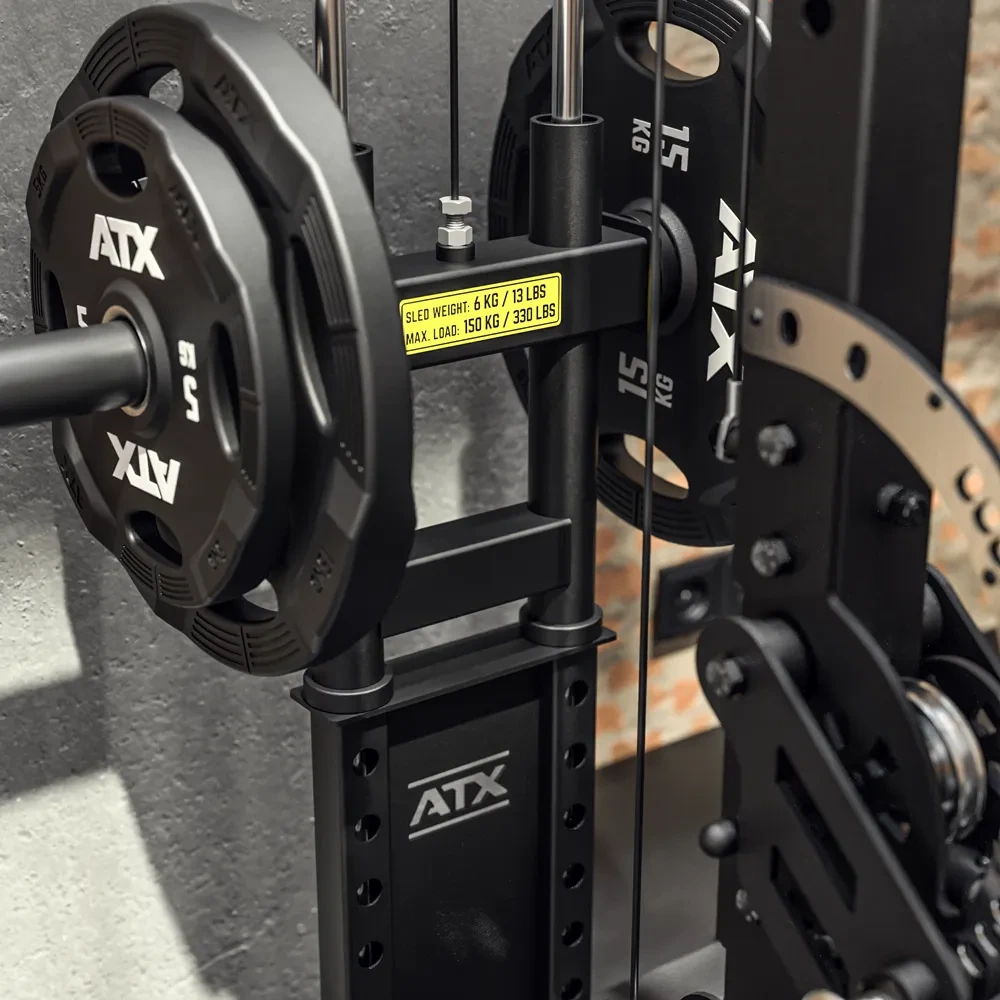 Close-up of a gym machine loaded with ATX-branded weight plates and a yellow label displaying weights and load information, set against a textured gray wall.