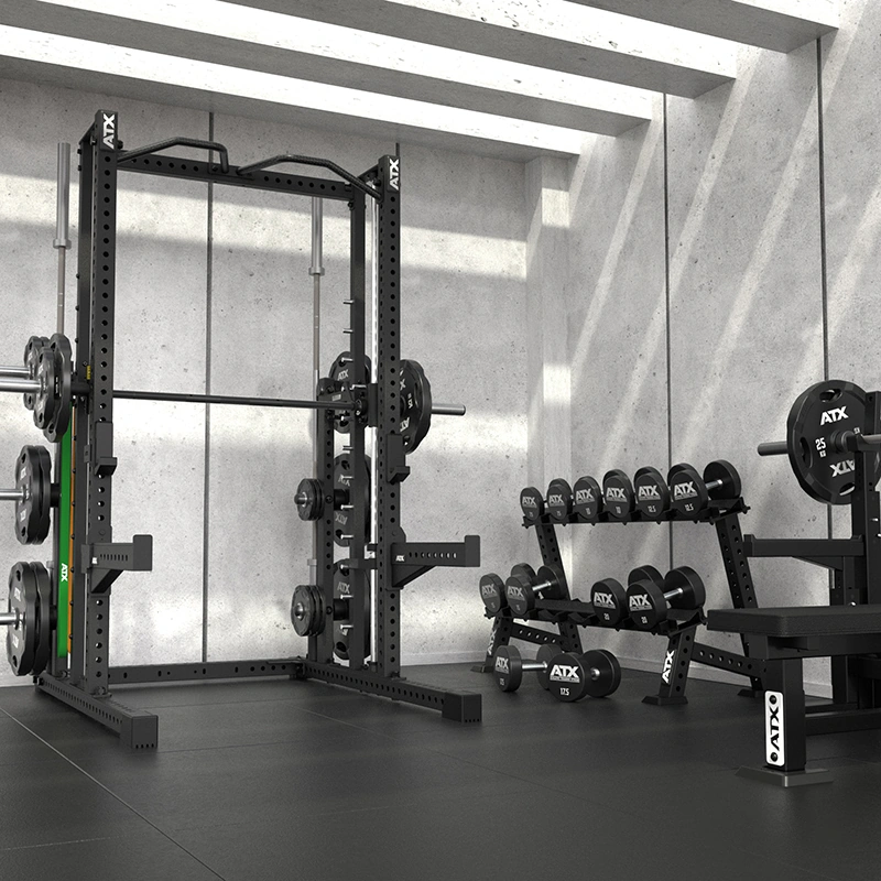 A modern gym featuring a squat rack, barbells, weight plates, and the ATX Commercial Smith Machine organized neatly next to dumbbells. Concrete walls and black flooring contribute to a minimalist, industrial aesthetic while natural light pours in from above.