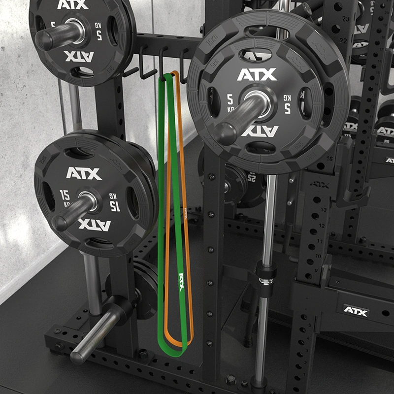 A gym rack with ATX weight plates, barbells, and two resistance bands—one green, one orange—hanging from the side stands alongside the ATX Commercial Smith Machine. The entire setup is placed against a concrete wall.