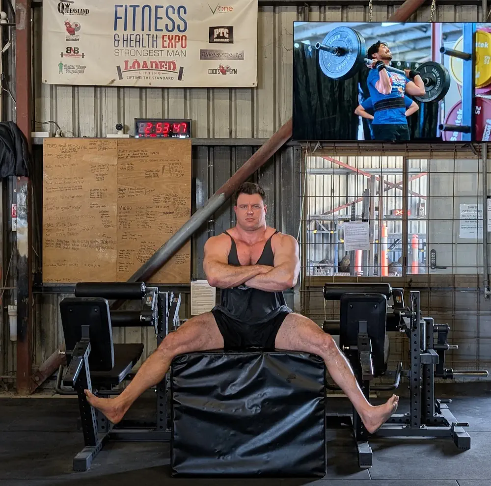 A muscular man in a gym sits on a padded platform with arms crossed and legs wide apart, perhaps fresh from his latest Seated Leg Extension Leg Curl session. Behind him are gym equipment, a digital clock, a large whiteboard, and a TV displaying a weightlifter in action.