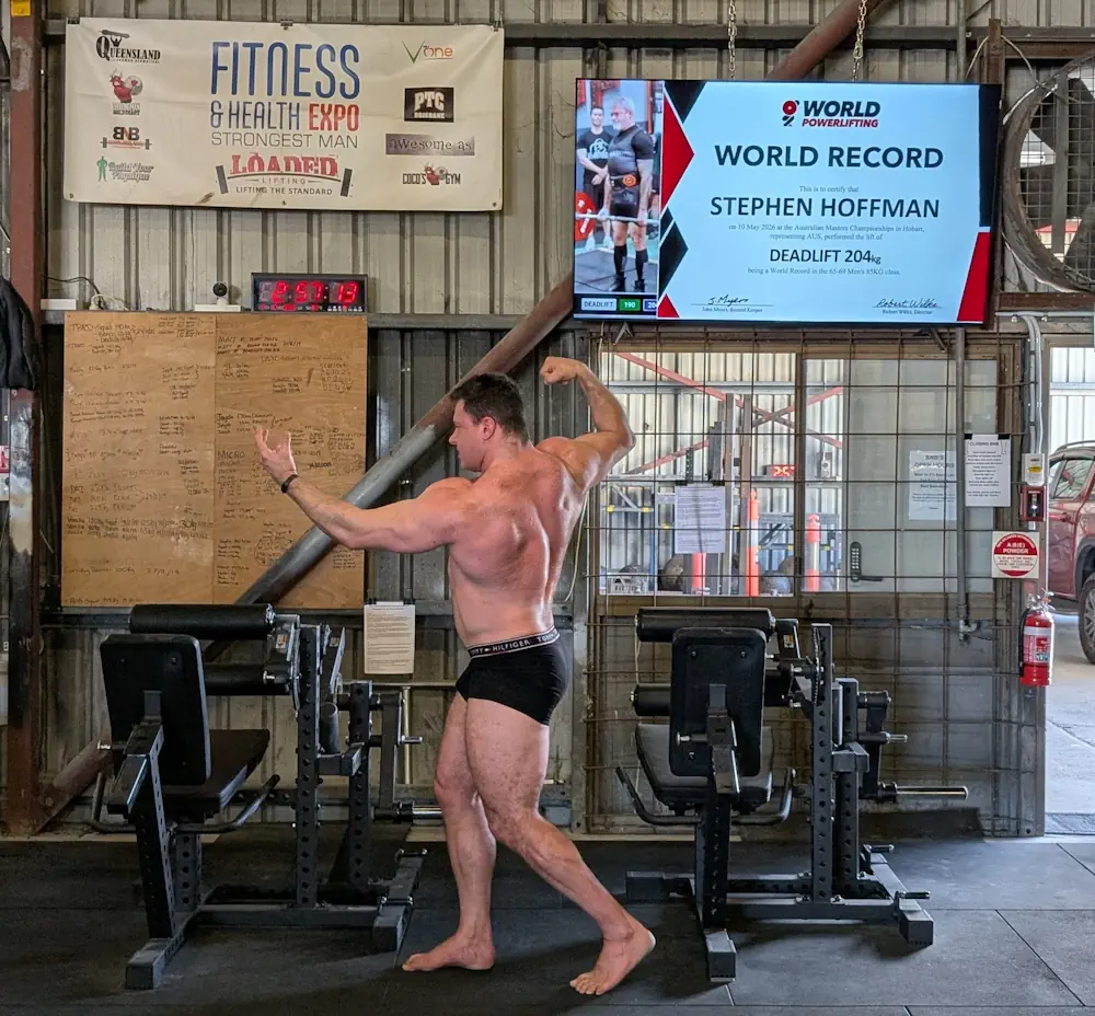 A muscular man in black shorts flexes in a gym. A screen behind him displays World Record for Stephen Hoffman, with a 204 kg deadlift. Nearby are chairs, a wall clock, and event banners, while a Seated Leg Extension Leg Curl Review poster adds to the athletic atmosphere.