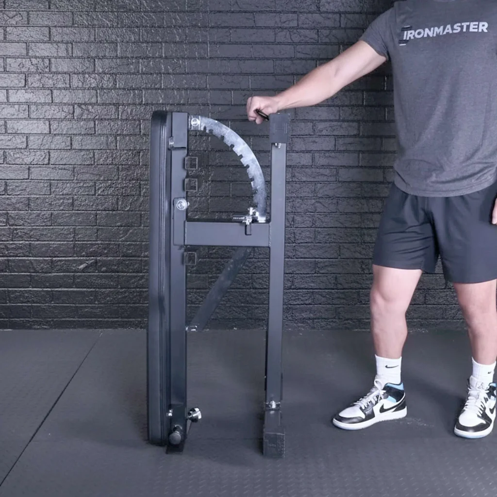 Wearing athletic apparel, a person stands beside a folded Super Bench equipped with the Kickstand for Super Bench, showcasing its compact storage against a black brick wall.