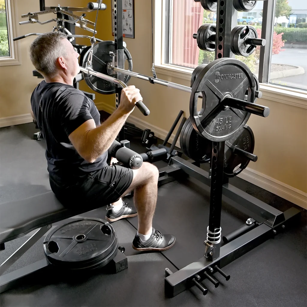 A man in black workout clothes performs a seated lat pulldown exercise using a barbell on a weight machine in a gym with mirrored walls and large windows.