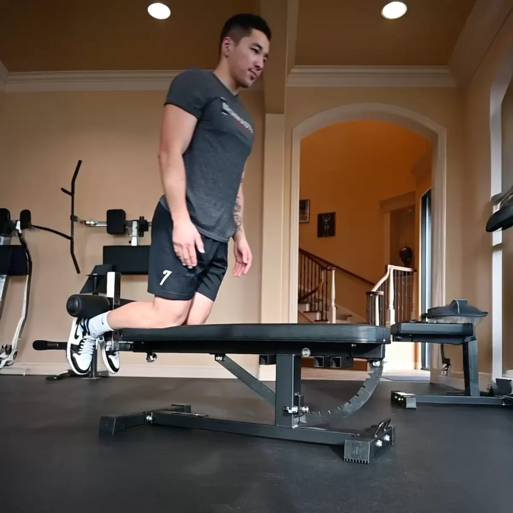 A man in athletic clothing kneels on a bench in a home gym, preparing to exercise. Fitness equipment, including a machine and weights, is visible in the background.