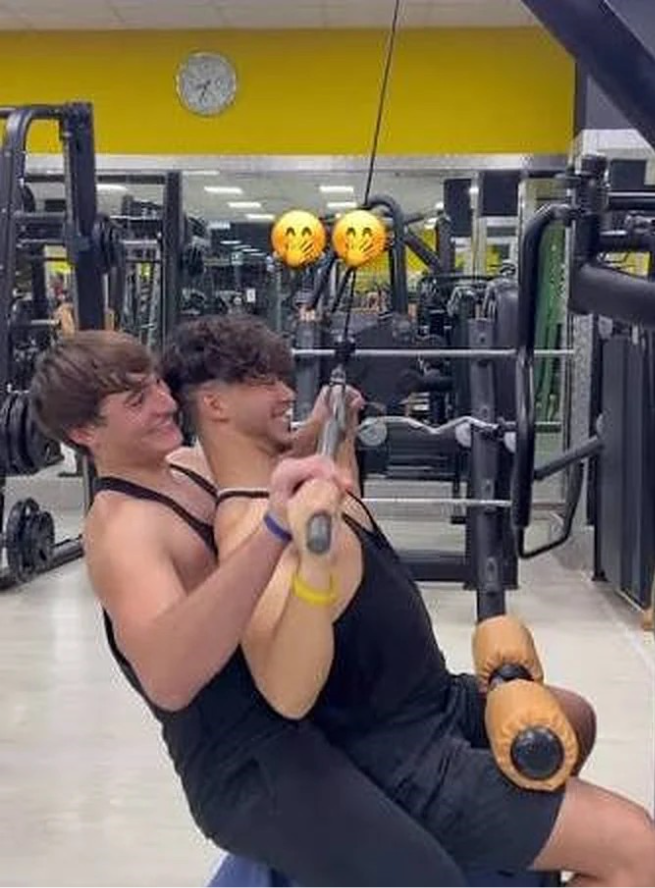 Two people sit back-to-chest on a gym machine, both pulling down on the handles and smiling. The faces in the mirror behind them are covered with emoji masks. Gym equipment and a clock are visible.