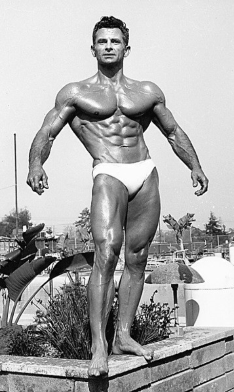 A male bodybuilder poses outdoors in a white posing suit, showing defined muscles. The background includes trees, plants, and what appears to be a pool area. The image is black and white.